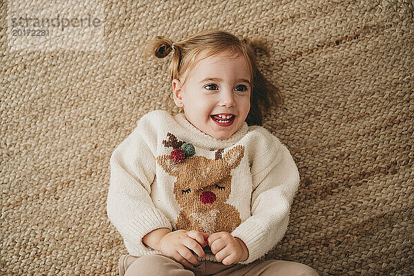 Lächelndes Mädchen trägt Weihnachtspullover und liegt zu Hause auf dem Teppich