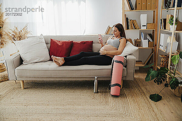 Schwangere Frau nutzt Smartphone und entspannt sich zu Hause auf dem Sofa