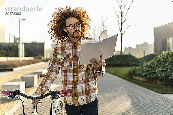 Glücklicher rothaariger Mann liest Buch neben Fahrrad auf Fußweg