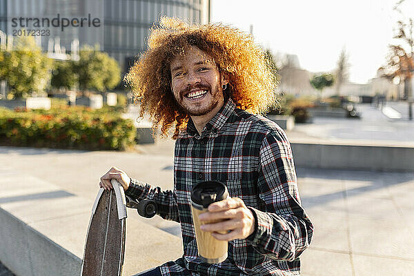 Lächelnder Mann sitzt an einem sonnigen Tag mit Kaffeetasse und Skateboard