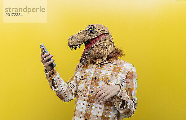 Mann mit Dinosauriermaske und Smartphone vor gelber Wand