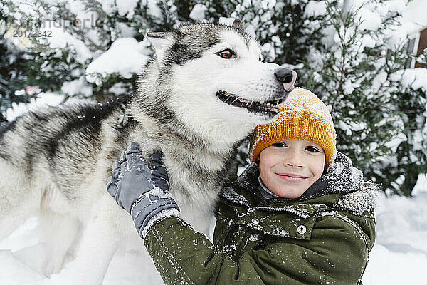 Lächelnder Junge streichelt Husky-Hund im Schnee neben Baum