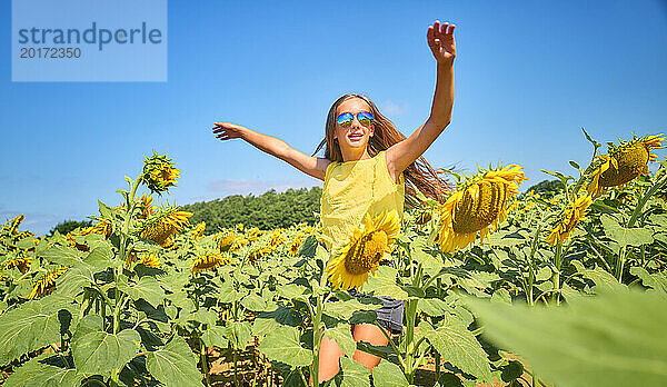 Fröhliches Mädchen mit Sonnenbrille und Tanz im Sonnenblumenfeld