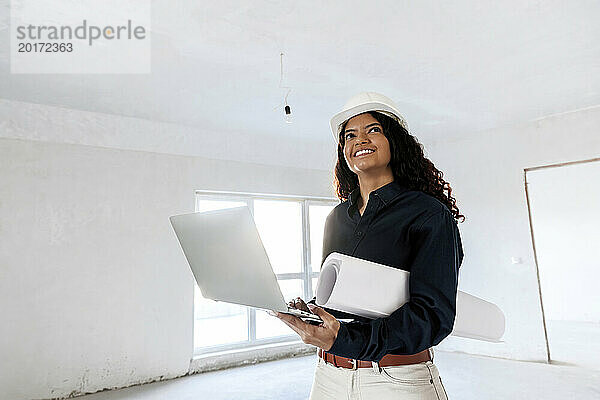 Glücklicher Architekt steht mit Bauplan und Laptop vor Ort