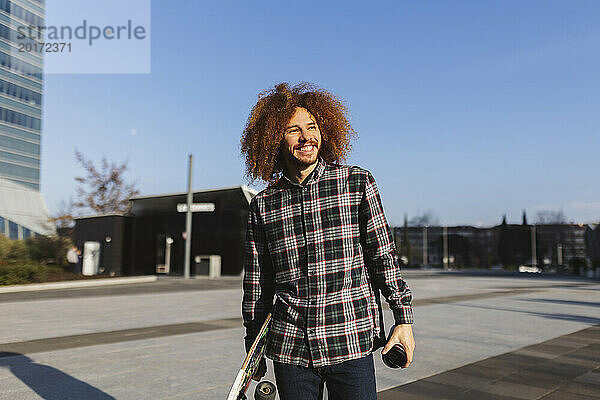 Glücklicher Mann mit rothaarigem lockigem Haar  der an einem sonnigen Tag Skateboard hält