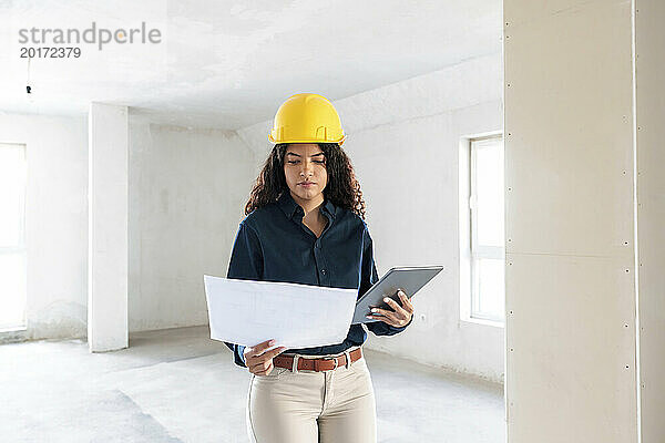 Architekt trägt gelben Schutzhelm und prüft Bauplan mit Tablet-PC vor Ort