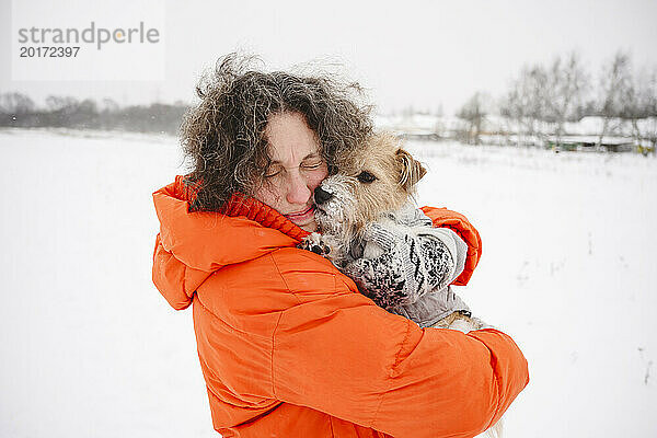 Frau trägt Winterjacke und umarmt Hund auf Schneefeld