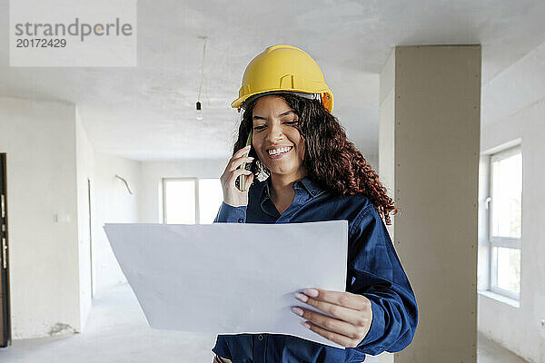 Glücklicher junger Architekt  der auf der Baustelle über den Bauplan auf dem Mobiltelefon diskutiert