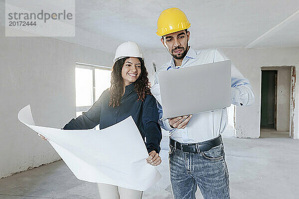 Lächelnder Architekt hält einen Bauplan in der Hand und diskutiert mit einem Kollegen vor Ort am Laptop