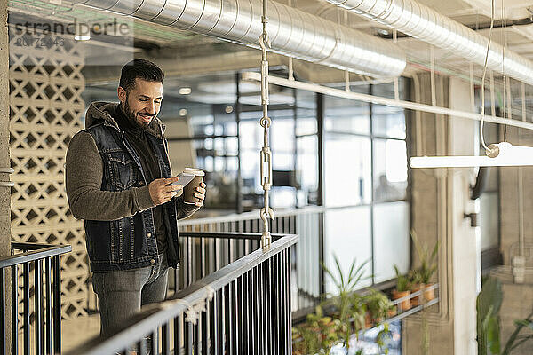 Lächelnder Geschäftsmann mit Smartphone und Einweg-Kaffeetasse im Büro