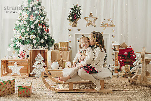 Mädchen küsst Schwester auf Schlitten neben Weihnachtsdekoration zu Hause