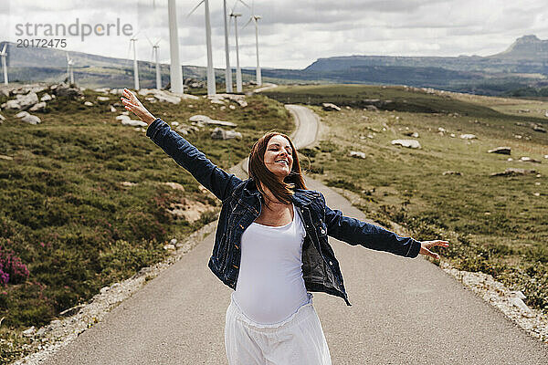 Spain  Madrid  Portrait of pregnant woman standing with raised arms in middle of road stretching past wind farm
