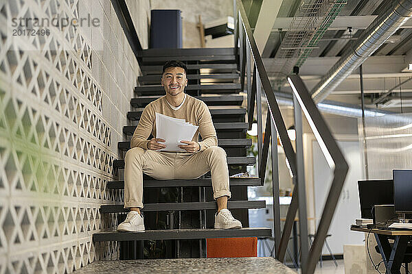 Lächelnder junger Geschäftsmann mit Dokumenten sitzt auf der Treppe im Büro