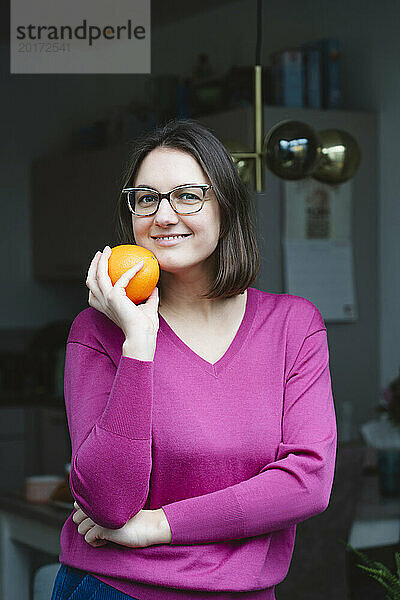 Lächelnde Frau hält Orange in der Hand und lehnt an der Haustür