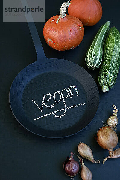 Studioaufnahme von Gemüse und schwarzer Bratpfanne mit dem Wort „vegan“ in Kreide geschrieben