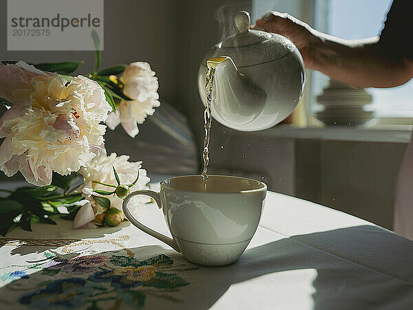 Frau gießt Tee in eine Tasse neben Blumen auf dem Tisch zu Hause