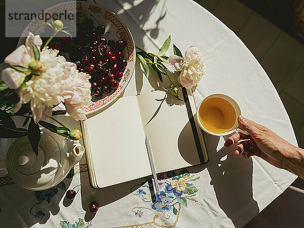 Hand einer Frau  die zu Hause eine Teetasse neben dem Tagebuch auf dem Tisch hält