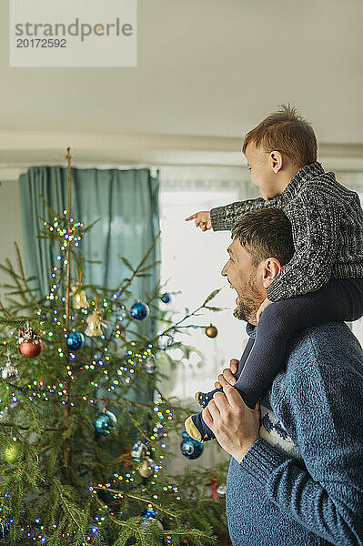 Fröhlicher Mann trägt Sohn auf Schultern neben dem Weihnachtsbaum zu Hause