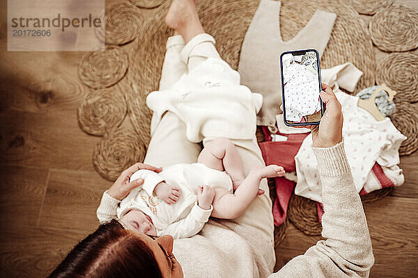 Frau hält Tochter und fotografiert Babykleidung mit Smartphone