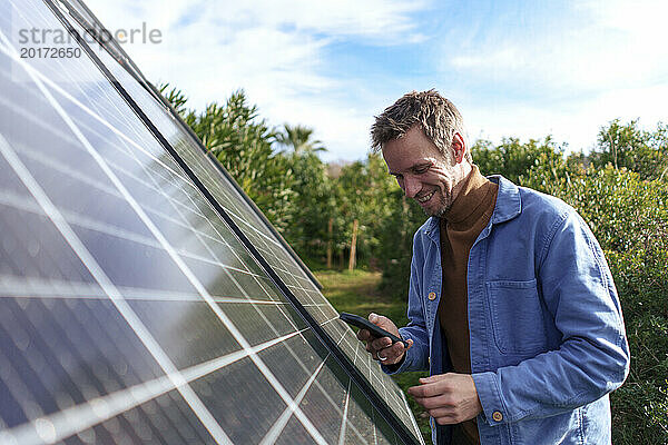 Glücklicher Mann  der an einem sonnigen Tag sein Smartphone in der Nähe von Sonnenkollektoren benutzt