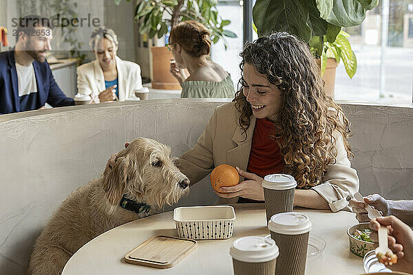 Glückliche junge Geschäftsfrau hält in der Cafeteria im Büro frische Orangenfrüchte neben dem Hund