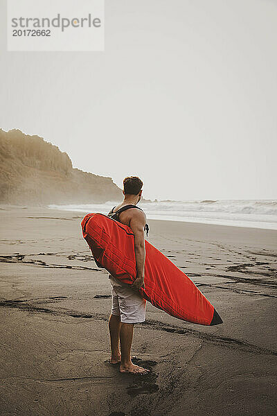 Junger Mann trägt Surfbrett und steht am Strand
