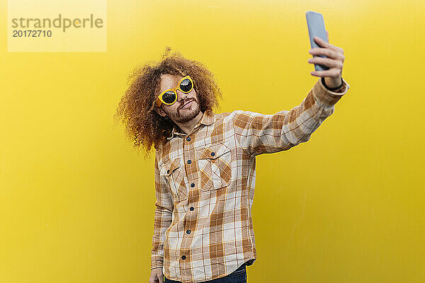 Mann trägt Sonnenbrille und macht Selfie mit Smartphone vor gelber Wand