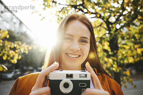 Smiling woman holding camera at sunny day in autumn