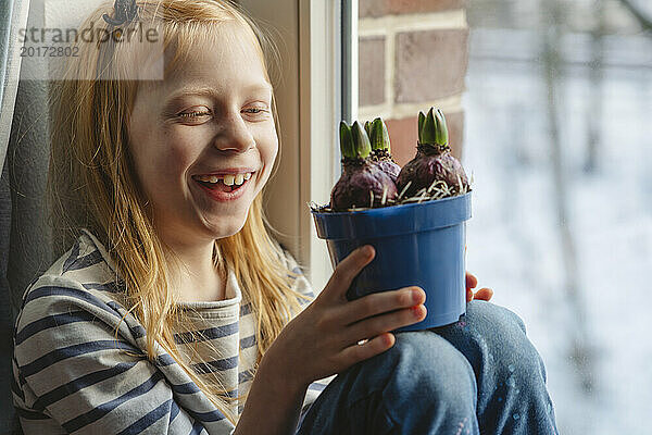 Glückliches Mädchen hält Hyazinthenzwiebeln in der Hand und sitzt am Fenster