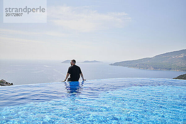Man looking at sea sitting on the edge of swimming pool at villa