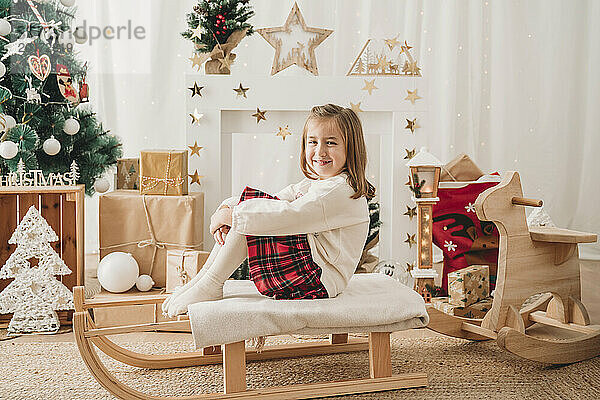 Lächelndes Mädchen sitzt auf Schlitten neben Weihnachtsdekoration zu Hause