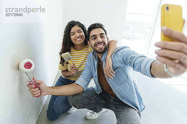 Verspieltes Paar mit Farbroller und Pinsel macht ein Selfie mit dem Smartphone im neuen Zuhause