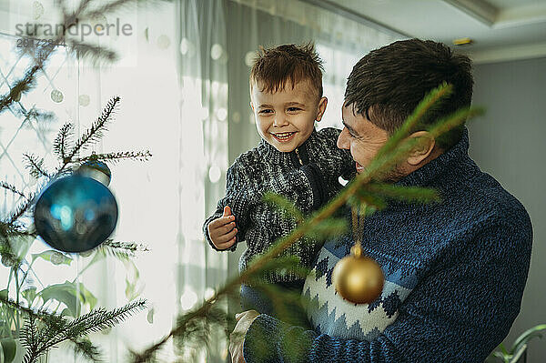 Glücklicher Vater trägt seinen Sohn zu Hause neben dem Weihnachtsbaum