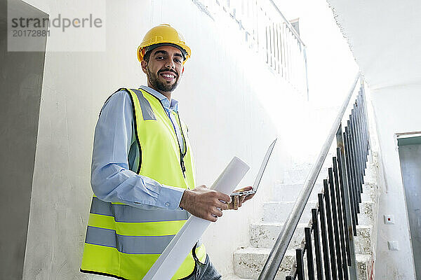 Lächelnder Architekt mit Schutzhelm steht auf Stufen und hält Laptop vor Ort