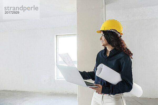 Lächelnder Architekt hält Laptop in der Hand und untersucht die Baustelle mit Bauplänen