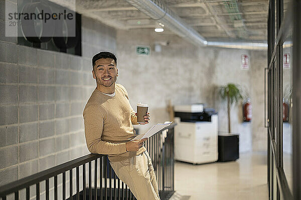 Lächelnder junger Geschäftsmann mit Einwegkaffeetasse und Dokumenten lehnt am Geländer im Büro