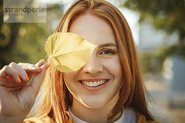 Lächelnde Frau bedeckt Auge mit Herbstblatt