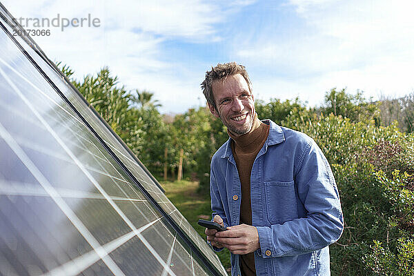 Glücklicher Mann  der an einem sonnigen Tag mit seinem Smartphone in der Nähe von Sonnenkollektoren steht