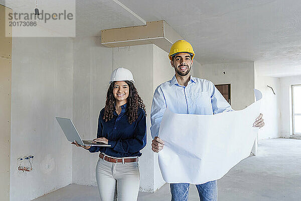 Lächelnde Architekten stehen mit Laptop und Bauplan vor Ort