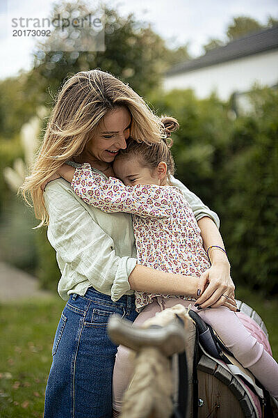 Mädchen sitzt auf Holzpferd und umarmt glückliche Mutter im Garten