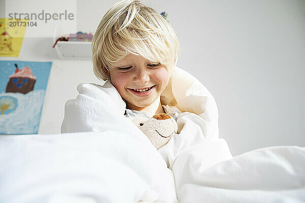 Glücklicher Junge  der zu Hause mit einem in eine Decke gehüllten Teddybär spielt