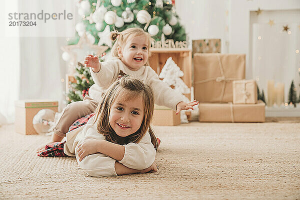 Glückliches Mädchen  das zu Hause auf dem Rücken der Schwester neben der Weihnachtsdekoration sitzt