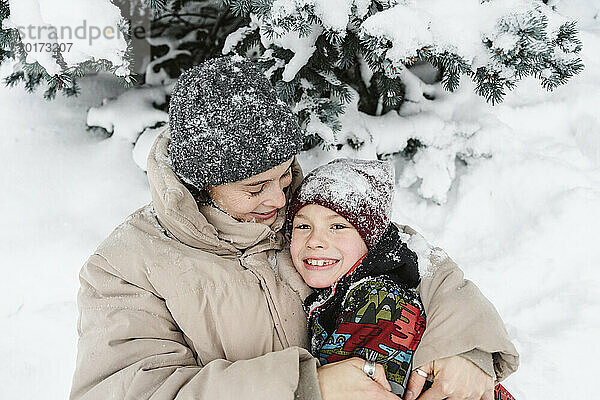 Glückliche Frau umarmt Sohn und liegt auf Schnee neben Baum