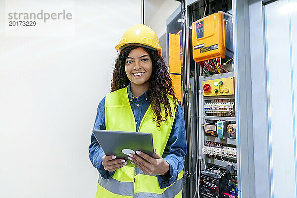 Lächelnder Ingenieur hält Tablet-PC am elektrischen Sicherungskasten