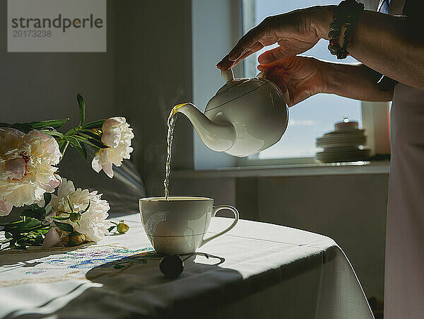 Reife Frau gießt zu Hause Tee in eine Tasse neben Blumen