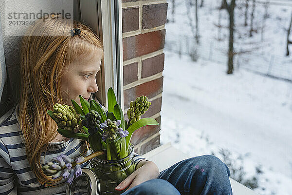 Mädchen hält einen Strauß Hyazinthenblüten im Glas und sitzt am Fenster