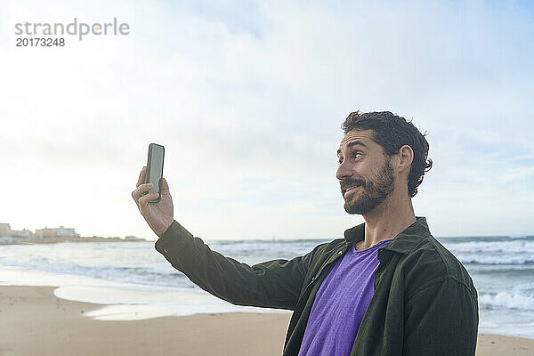Lächelnder Mann macht ein Selfie mit seinem Smartphone vor dem Meer am Strand