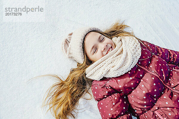 Glückliches blondes Mädchen  das sich mit geschlossenen Augen auf den Schnee legt