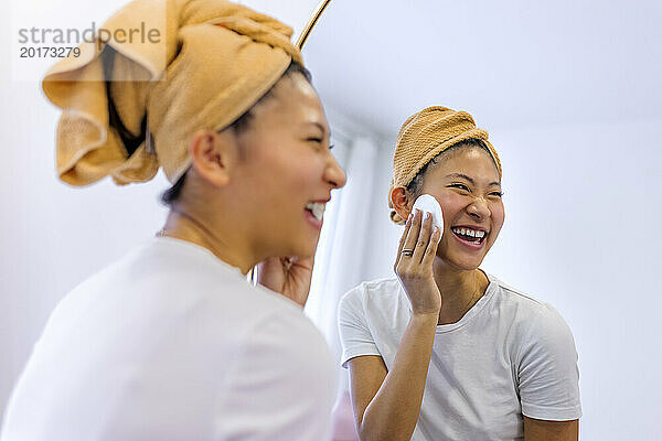 Fröhliche Frau reinigt ihr Gesicht mit Wattestäbchen vor dem Spiegel zu Hause