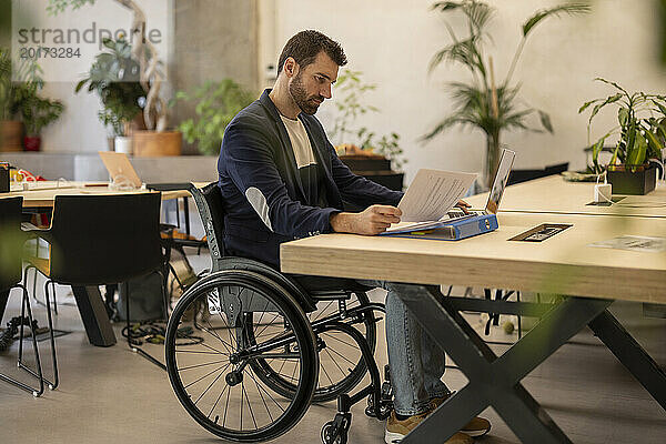 Selbstbewusster Geschäftsmann im Rollstuhl mit Behinderung prüft Dokument am Schreibtisch